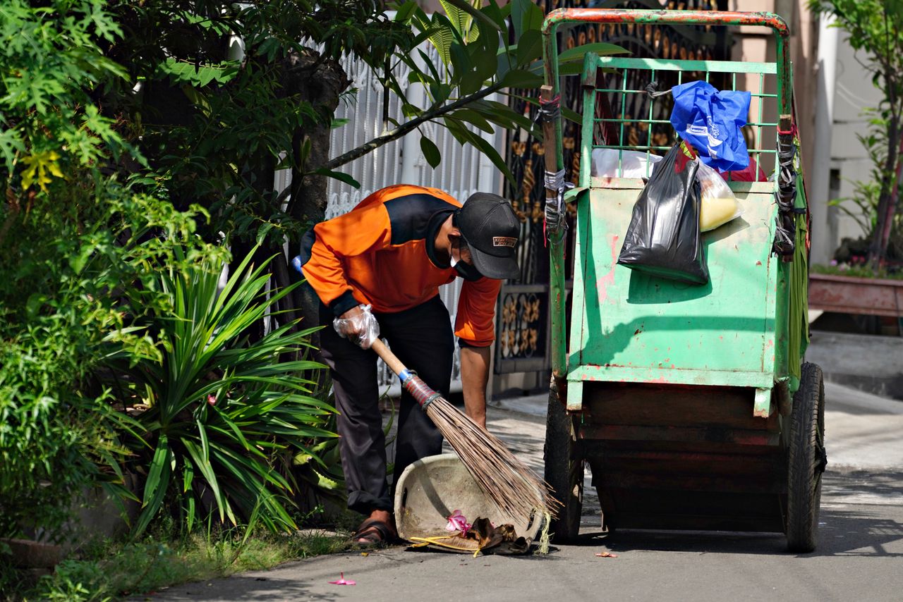 A street sweeper in uniform cleans a city sidewalk using a broom and cart, emphasizing urban cleanliness.